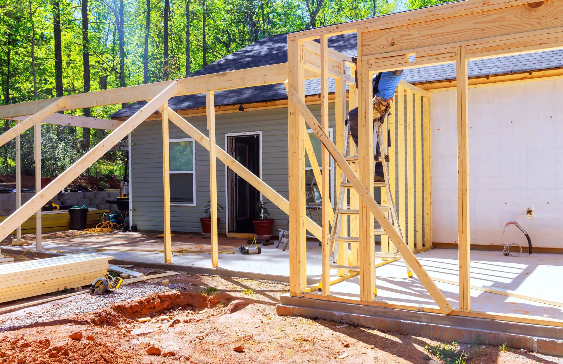 Timber framing for a ground-floor addition standing on a poured concrete slab beside a house with pale green horizontal siding and a dark shingled roof, with a person on a stepladder working at the top plate, stacked lumber, a circular saw and coiled extension cords on the ground, and trees behind.