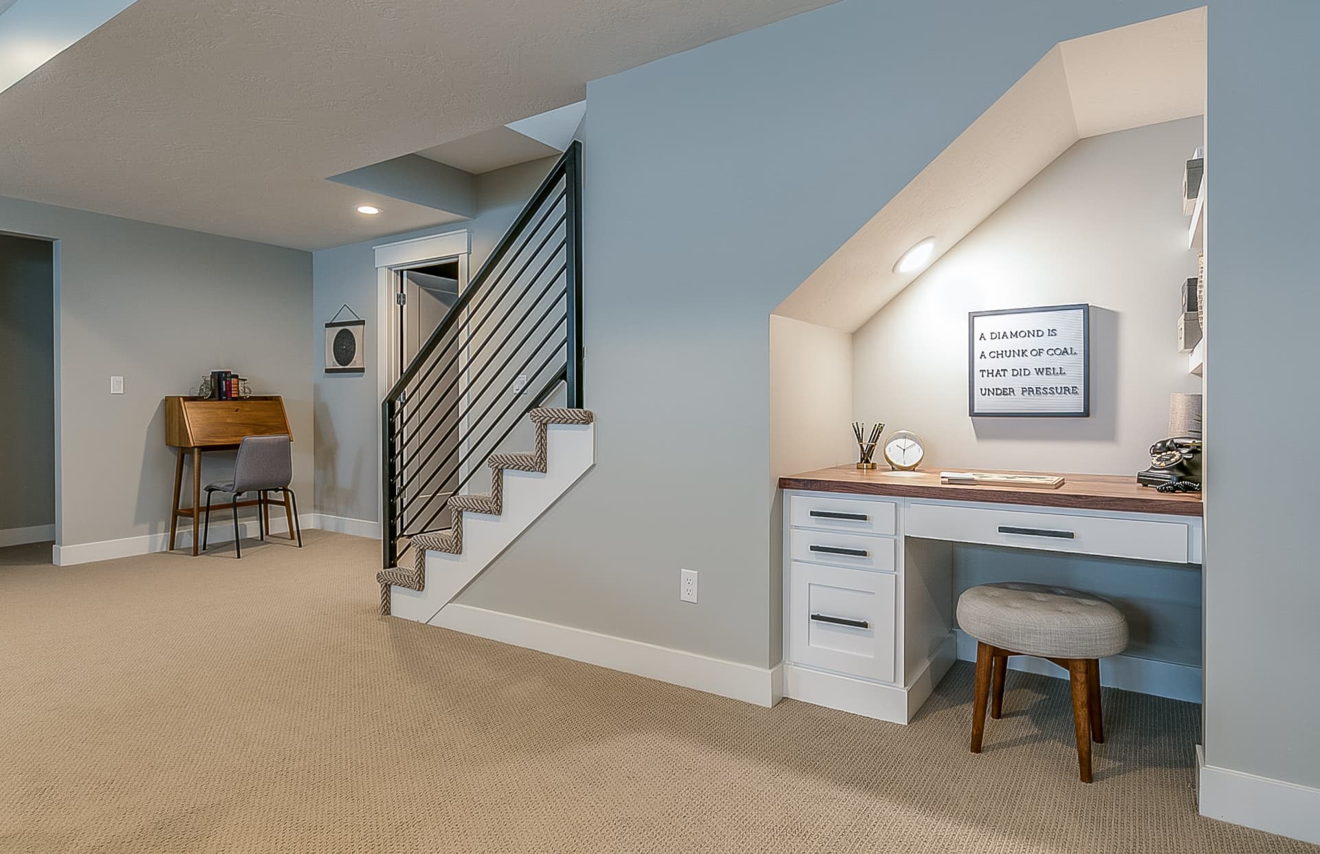 A finished lower-floor living area with pale blue walls, carpeted floor, a staircase behind a black horizontal railing, and a built-in desk with white drawers set into a sloped-ceiling alcove.