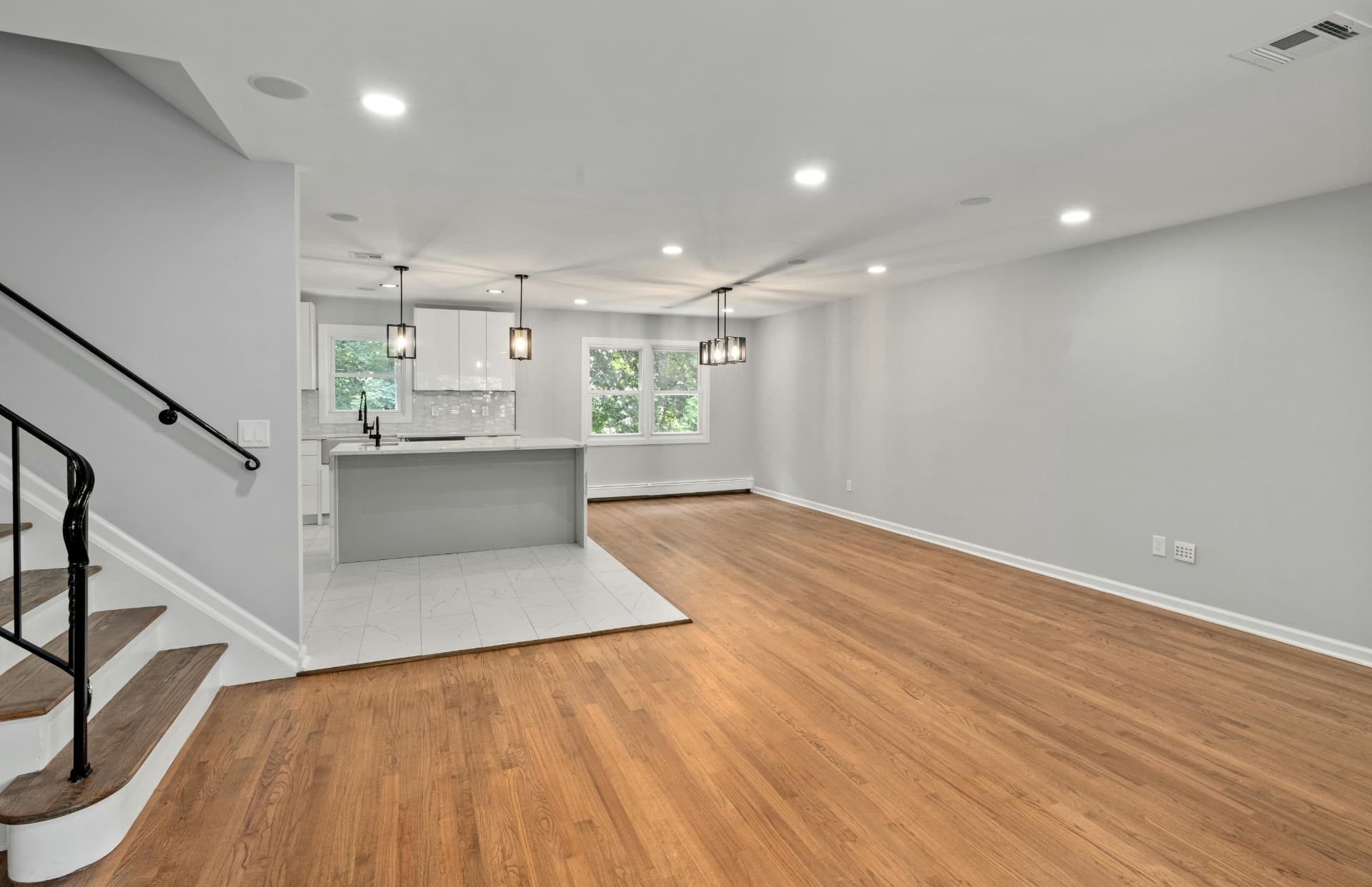 A finished open-plan lower floor with oak flooring, grey walls, recessed and pendant lighting, and a compact kitchenette with white cabinets, a stone-topped island and windows looking onto trees.