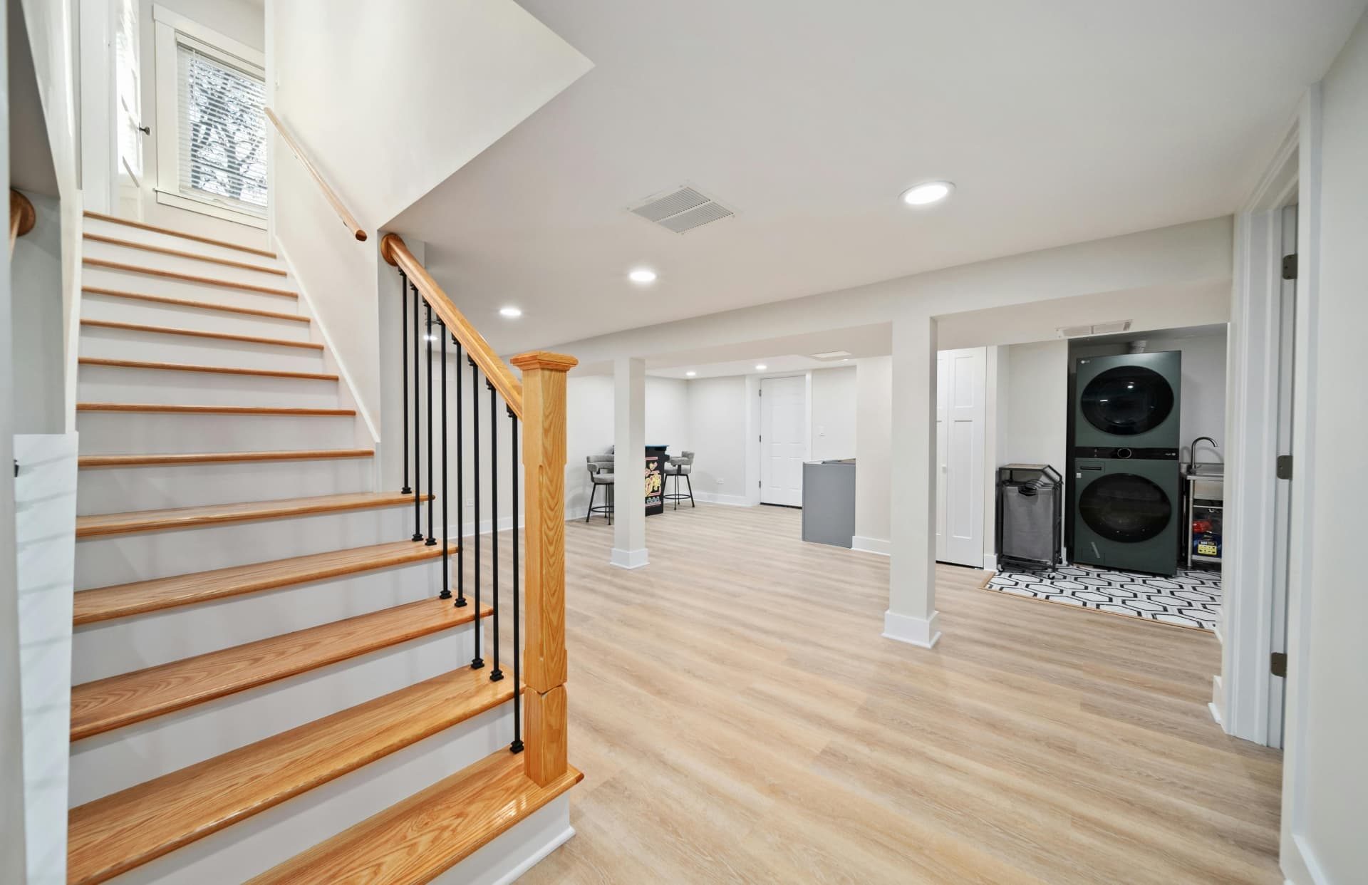 A finished lower floor with pale walls and light wood-look flooring, an oak staircase with black metal spindles in the foreground, recessed ceiling lights, and a stacked washer and dryer in an open laundry area beyond.