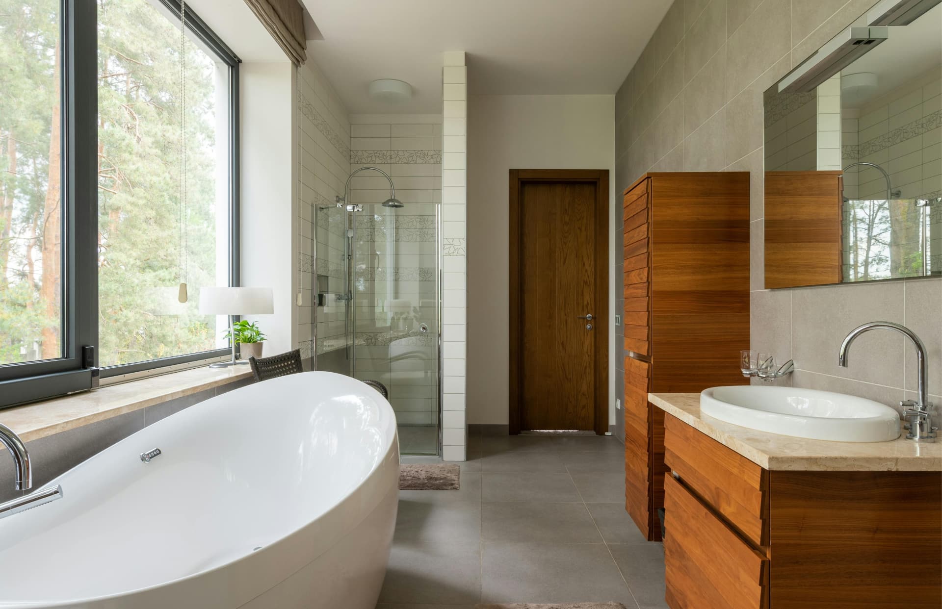 A finished bathroom with a freestanding oval bath beside a full-height window, a glass shower enclosure, tiled walls and floor, and a wood vanity with a round basin below a wall mirror.