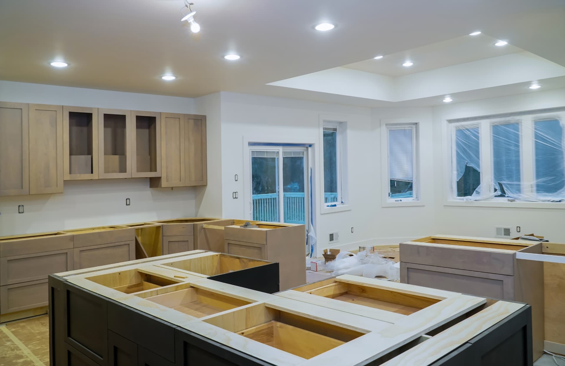 A kitchen part-way through installation, with wall cabinets hung and base cabinet boxes and a large island carcass standing open without countertops or doors, recessed ceiling lights on and the windows masked with protective plastic.